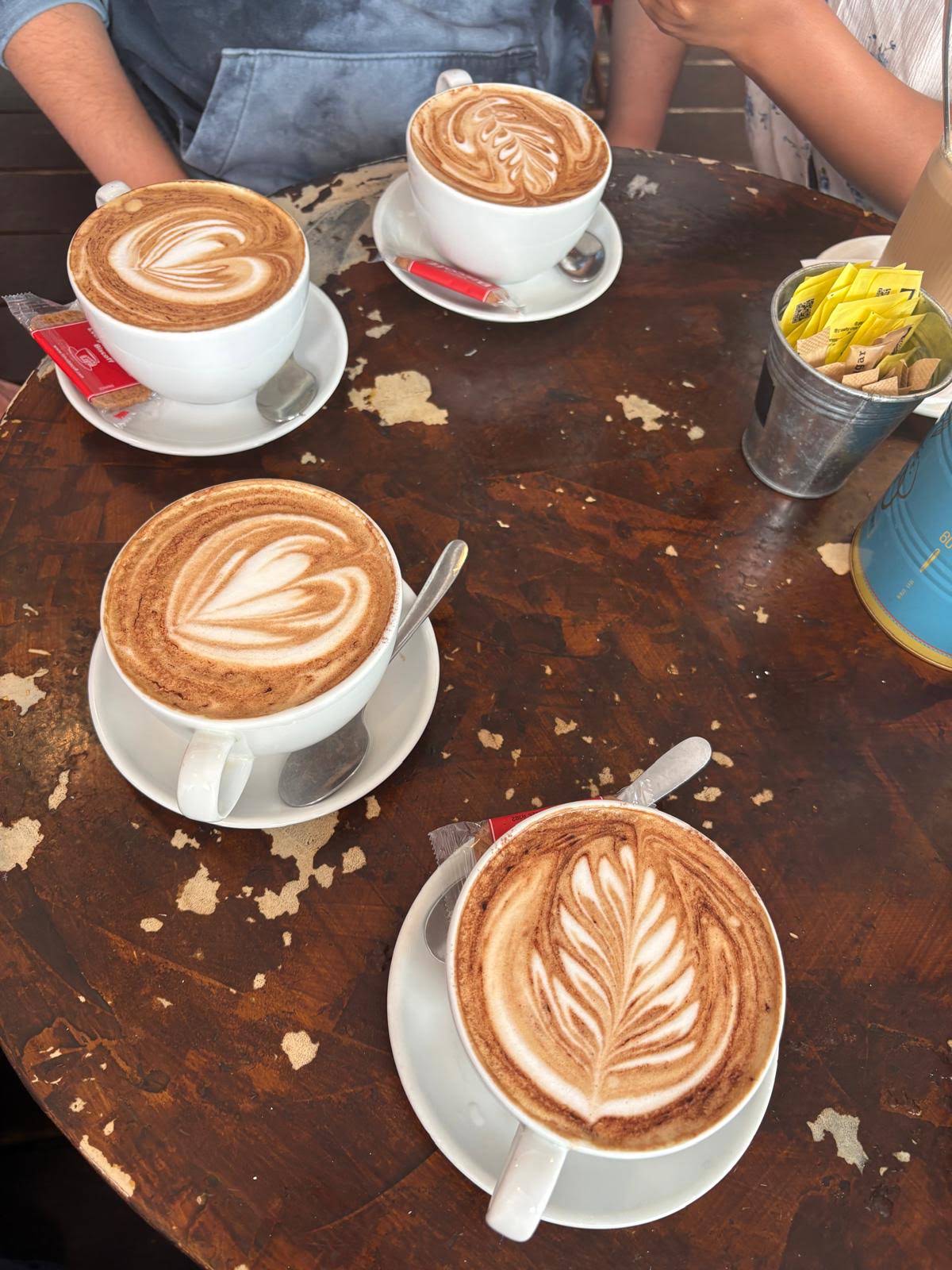 Four complete coffee cups and saucers with leaf-pattern foam on a worn wooden table at the George Street coffeehouse
